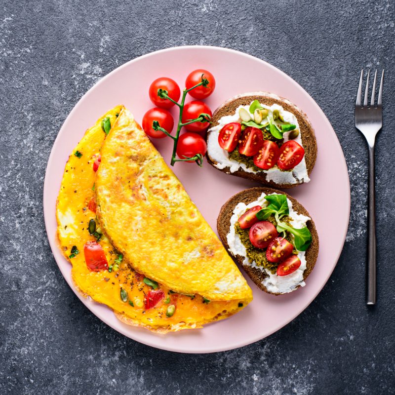 top view healthy Breakfast of egg omelet, wholegrain toast with cream cheese, pesto and cherry tomatoes on pink plate, on black stone background with copy space.
